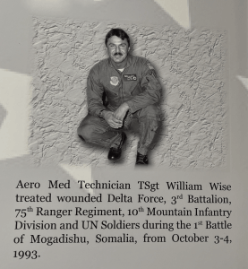 Image of Bill Wise, an aeromedical evacuation crew member, in a flight suit. Wise sports a chevron-style mustache slightly wider than his lips. On his right breast of his flight suit is the Military Airlift Command. His black name plate has enlisted wings and the bucking cowboy emblem of Wyoming. 