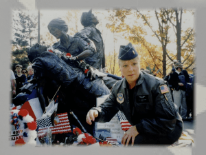 Image of Lee Carruthers, a blonde flight nurse in a green flight suit, green jacket, and boue garrson cap with a lieutenan's bar. She's kneeling down in front of the Vietnam Women's memorial in Washington, D C.